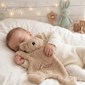 Peut inclure: Un bébé endormi serre contre lui une couverture de sécurité en forme d'ours en peluche beige, brodée du nom "Liam". Le bébé porte un pyjama crème. Un lapin crocheté et des blocs de bois sont en arrière-plan.