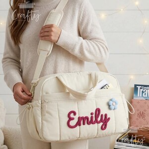 Puede incluir: Bolso cambiador acolchado color crema con correa para el hombro y asas superiores. La bolsa presenta el nombre "Emily" en fuente cursiva y una aplicaci&oacute;n de flor azul. Tiene m&uacute;ltiples bolsillos y cierre de cremallera.