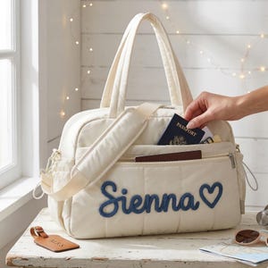 Puede incluir: Bolso de viaje acolchado color crema con el nombre "Sienna" y un coraz&oacute;n bordados en azul. El bolso tiene una correa para el hombro, asas superiores y m&uacute;ltiples bolsillos. Se est&aacute; colocando un pasaporte en el bolsillo delantero. Una etiqueta de equipaje es visible.