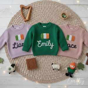 May include: Three children's sweaters in lavender, green, and pink, each with an embroidered Irish flag and a name. The sweaters are displayed on a beige crocheted mat, with decorative items like a harp, sheep, and a leprechaun.