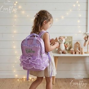 May include: A lavender backpack featuring multiple layers of ruffled tulle fabric, a small decorative bow, and the name Chloe embroidered in green thread on the front pocket. The bag includes two top handles and adjustable shoulder straps.