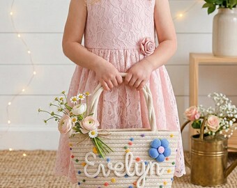 Puede incluir: Una cesta de cuerda beige con el nombre "Evelyn" en escritura blanca, decorada con pompones coloridos y una flor azul. La cesta está llena de flores y la sostiene una persona que lleva un vestido de encaje rosa.