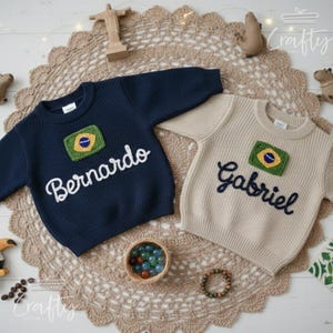 May include: Two children's sweaters, one navy blue and one beige, are displayed on a decorative surface. Each sweater features a Brazilian flag patch and a name embroidered in cursive: "Bernardo" and "Gabriel".