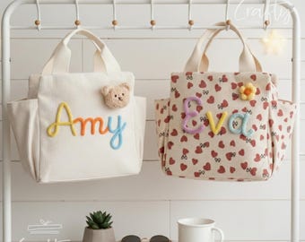 Puede incluir: Dos bolsas de mano de lona. Una es de color blanco roto con el nombre "Amy" en bordado colorido y un aplique de oso de peluche. La otra tiene un estampado de corazones rojos y el nombre "Eva" en bordado colorido, con un aplique de flor.
