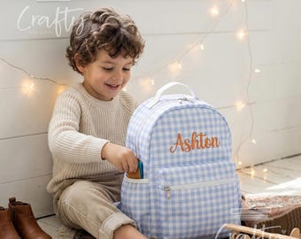 Puede incluir: Una mochila de cuadros vichy en azul claro y blanco con un asa superior blanca, un bolsillo frontal con cremallera y el nombre Ashton bordado en letra cursiva naranja en la parte delantera. Incluye un bolsillo lateral y cremalleras plateadas.