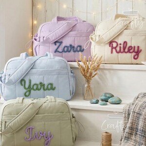 Puede incluir: Cuatro bolsas de beb&eacute; acolchadas en colores pastel: lavanda, azul claro, crema y verde salvia. Cada bolsa tiene un nombre bordado en un color contrastante: "Zara", "Yara", "Riley" e "Ivy". Las bolsas tienen asas superiores y correas para el hombro.