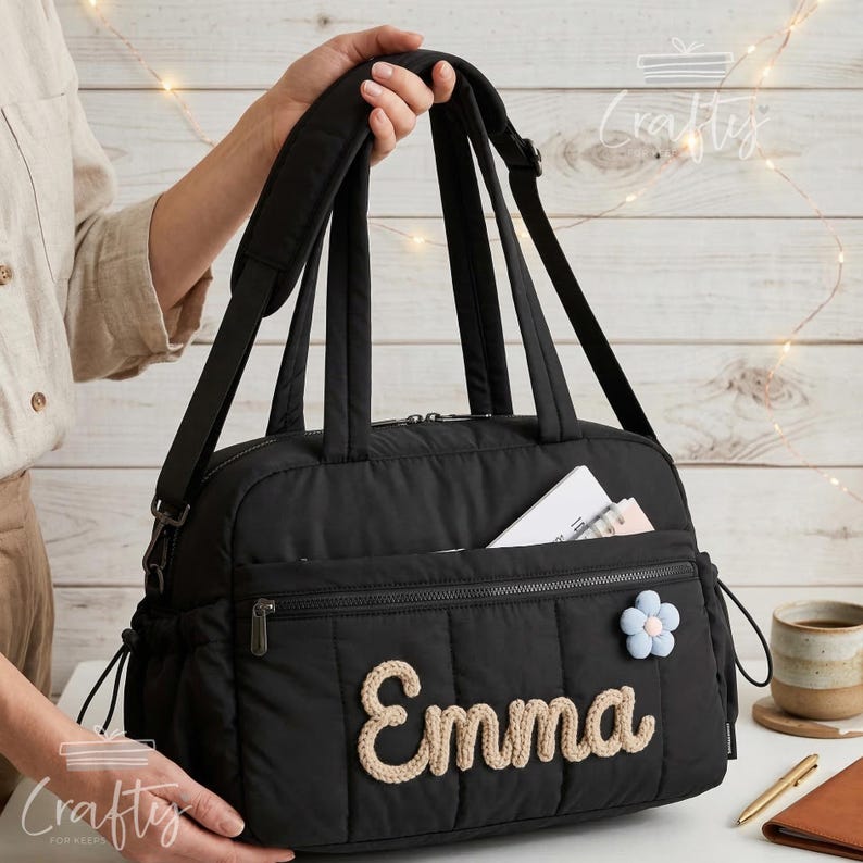 Puede incluir: Bolso tote acolchado negro con un bolsillo con cremallera y un adorno floral azul claro y rosa. El nombre "Emma" est&aacute; bordado en hilo color crema. El bolso tiene una correa para el hombro y dos asas.