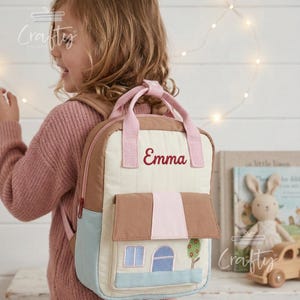 May include: A beige and brown backpack with a house design, featuring a blue base, brown roof, and a pink accent. The name "Emma" is embroidered in red. The backpack has pink straps and a handle.
