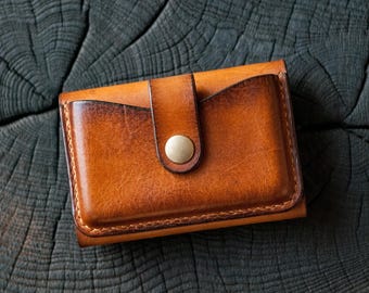 May include: A handcrafted, brown leather wallet with a button closure. The wallet has a rich, warm color with darker edges, suggesting a natural, aged finish. The design includes card slots and a secure flap.
