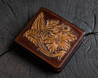 Pode incluir: Uma carteira de couro castanha com um design floral em relevo detalhado na capa frontal. A superfície apresenta um padrão esculpido de uma grande flor central rodeada por folhas e vinhas, com um acabamento em castanho escuro.