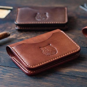 May include: Close-up of several brown leather cardholders with a cat design embossed on the front. The cardholders are rectangular with rounded corners and visible stitching. The leather has a rich, warm tone.