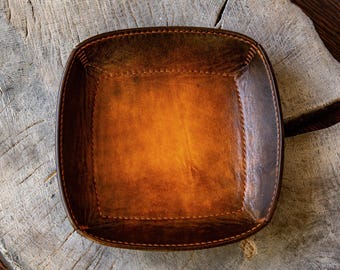 May include: A square-shaped valet tray crafted from brown leather with a distressed, multi-tonal finish. The tray features raised, folded edges secured with visible, thick stitching along the perimeter, creating a deep, open-top container.
