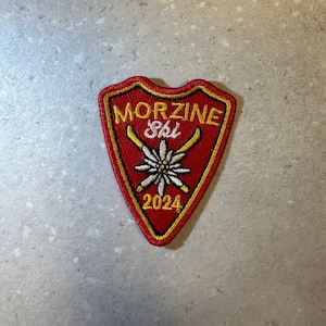 Puede incluir: Parche bordado rojo y dorado con las palabras "MORZINE Ski 2024" y un diseño floral. El parche presenta esquís cruzados y forma de escudo, perfecto para los amantes del esquí.