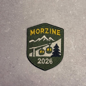 Puede incluir: Parche bordado con forma de escudo, con la palabra "MORZINE" en amarillo, encima de una cordillera y un diseño de telesilla. El año "2026" está debajo del telesilla. El parche tiene un borde y un fondo verde oscuro.