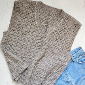 May include: A light brown, hand-crocheted sweater with a V-neck design. The sweater has long sleeves and a textured stitch pattern. A pair of blue denim jeans is partially visible in the bottom right corner, suggesting a casual outfit.