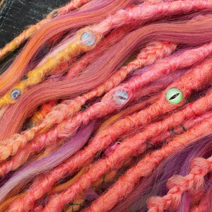 May include: A close-up of a set of colorful synthetic dreadlocks with beads. The dreadlocks are a mix of pink, orange, and purple.
