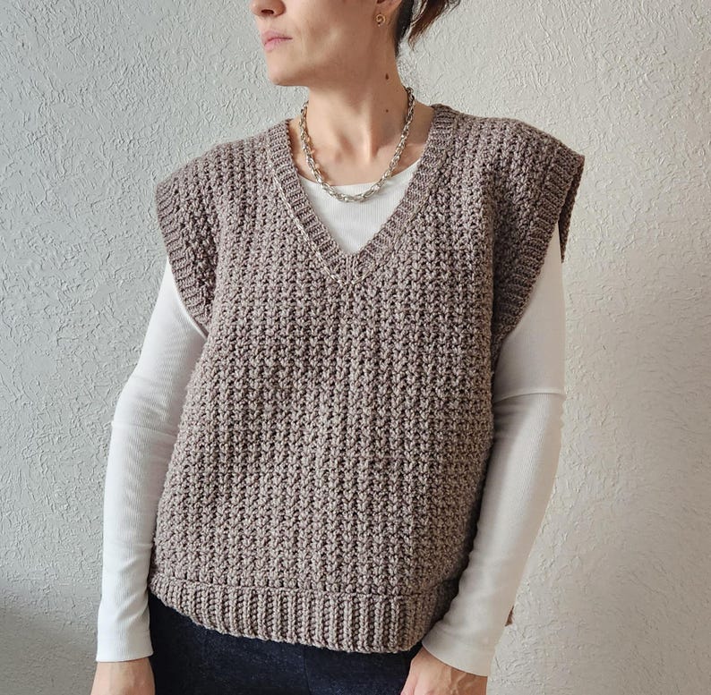 May include: A brown, textured, V-neck sweater vest. The vest is made of a knitted material and has a ribbed hem. The person is wearing a white long-sleeve shirt underneath and a silver chain necklace.