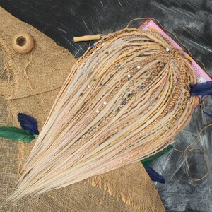 May include: Blonde and light brown dreadlocks with decorative beads and accents. The dreadlocks are arranged in a fan shape, with a mix of colors and textures. The background includes burlap fabric, feathers, and a dark surface.