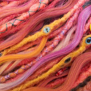 May include: Close-up of colorful synthetic dreadlocks with beads and charms. The dreadlocks are in shades of pink, orange, yellow, and purple.