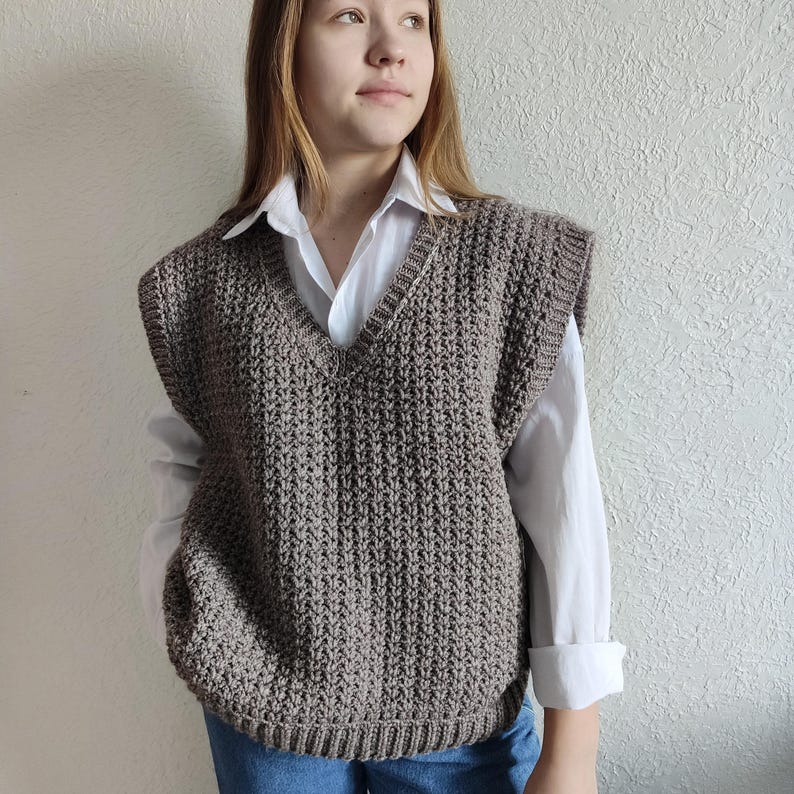 May include: A brown, knitted V-neck vest. The vest is sleeveless and has a textured stitch pattern. The person is wearing a white collared shirt underneath. The vest is a neutral color and appears to be handmade.
