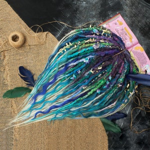 May include: A set of handmade dreadlocks in shades of blue, green, and purple, adorned with decorative gold leaves and butterfly accents. The dreadlocks are arranged on a burlap surface with a book in the background.