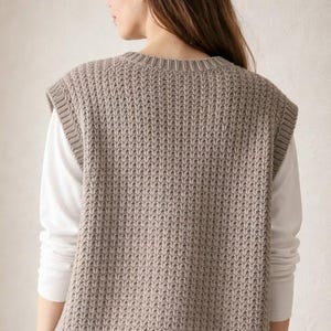 May include: A neutral-toned, knitted vest with a textured pattern. The sleeveless garment has a crew neck and a ribbed hem. The vest is worn over a white long-sleeved shirt. The overall style is casual and comfortable.