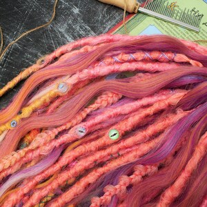 May include: A bundle of synthetic dreadlocks in shades of pink, orange, and yellow. The dreadlocks are made of kanekalon hair and are approximately 24 inches long.