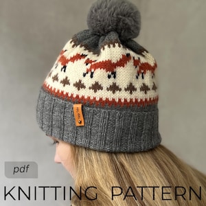 Può includere: Un cappello di lana grigio con un motivo a volpe arancione, bianco e marrone. Il cappello ha un pompon grigio e un'etichetta in pelle con la scritta "Triple Tribe". Il testo "KNITTING PATTERN" è visibile in basso.
