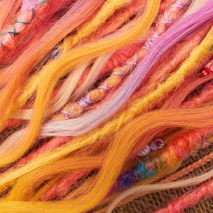 May include: A close-up of a variety of colorful synthetic hair extensions. The strands are woven together in a loose braid, creating a textured and vibrant look. The colors include shades of orange, yellow, pink, and purple.