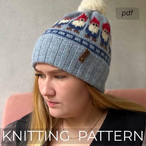 Op de afbeelding: Een lichtblauwe gebreide muts met een crèmekleurige pompon. De hoed heeft een herhalend patroon van kabouters met rode hoeden en blauwe jassen. De tekst "KNITTING PATTERN" staat onderaan de afbeelding.
