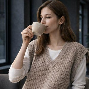 May include: A woman wearing a beige knitted vest over a white long-sleeved shirt, holding a beige mug to her lips. The vest has a V-neck and a textured knit pattern. The background is blurred, suggesting an indoor setting.