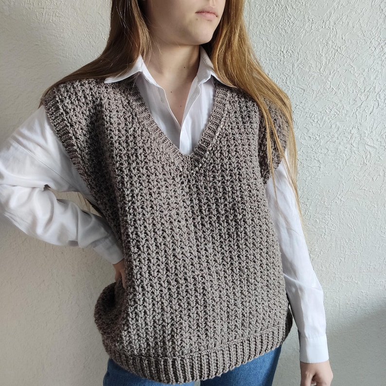 May include: A taupe-colored, knitted V-neck vest. The vest has a textured stitch pattern and ribbed hem and neckline. The vest is worn over a white collared shirt. The overall style is casual and comfortable.