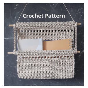 May include: A beige crochet wall organiser with wooden dowels and the text "Crochet Pattern". The organiser has two compartments and is hanging on a wall. It contains a white folder and a tan notebook.