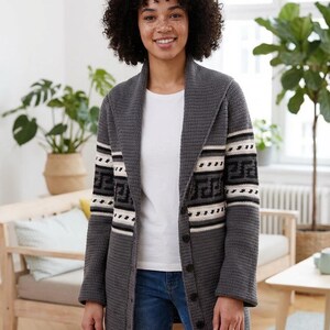 May include: A gray crochet cardigan with a shawl collar and button closure. The sleeves feature a geometric pattern in black, white, and beige. The cardigan is worn over a white t-shirt and blue jeans.