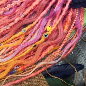 May include: A close-up of a bundle of colorful synthetic dreadlocks with pink, yellow, and white strands. The dreadlocks are decorated with small beads and charms.