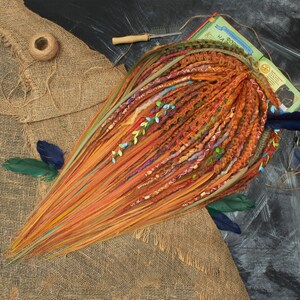 May include: A collection of orange and brown dreadlocks, adorned with colorful accents and decorative elements. The dreadlocks are arranged on a burlap surface, with a book and other accessories in the background. The overall aesthetic is bohemian and artistic.
