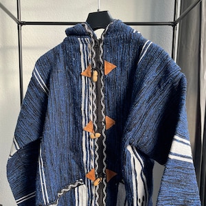 Op de afbeelding: Een blauwe en witte gestreepte capuchontrui met bruine leren knopen en een ritssluiting. De trui heeft een geweven patroon en een losse pasvorm.