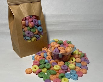 May include: A brown paper bag with a clear window showing colorful cereal rings. A small white cup filled with the same cereal rings is in front of the bag. The cereal rings are pink, blue, yellow, and green.