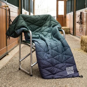 Könnte beinhalten: Eine gesteppte Decke in Grün- und Blautönen ist über einen silbernen Klappstuhl drapiert. Die Decke hat ein Rautenmuster und der Text "HORSE SHOW MOM" ist in der unteren Ecke aufgedruckt. Der Stuhl steht in einem Stall.
