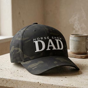 May include: A black camouflage baseball cap with the words "HORSE SHOW DAD" in white block letters. The cap has a curved brim and a button on top. The background includes a mug and a neutral-toned surface.