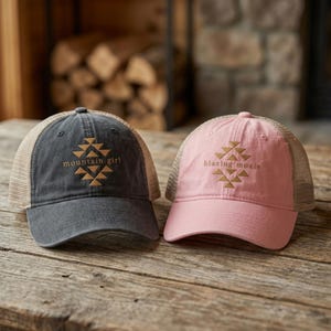 Könnte beinhalten: Eine anthrazitfarbene und eine hellrosa Trucker-Cap, jeweils mit beigem Netzrücken und einem beigefarbenen, gestickten geometrischen Rautenmuster. Die anthrazitfarbene Cap zeigt den Text mountain girl, die rosa Cap zeigt blazing moxie.