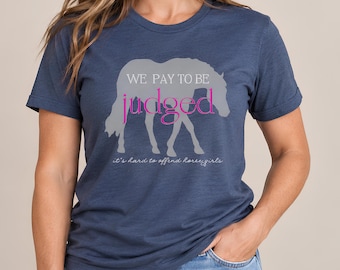 Könnte beinhalten: Ein kurzärmeliges T-Shirt in meliertem Marineblau mit einer hellgrauen Pferdesilhouette. Der Text lautet WE PAY TO BE judged in Weiß und Pink, darunter steht der kleinere weiße Schreibschrifttext it's hard to offend horse girls.