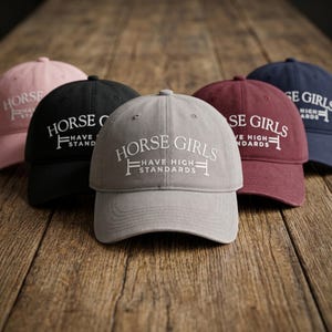 Può includere: Cinque cappellini da baseball in vari colori, tra cui rosa, nero, grigio, bordeaux e blu navy, sono esposti su una superficie di legno. Ogni cappellino presenta il testo bianco "HORSE GIRLS HAVE HIGH STANDARDS".