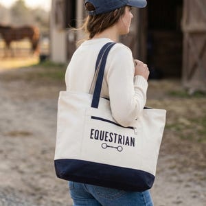 May include: Cream canvas tote bag with navy blue detailing. The bag has a zipped pocket and navy blue handles. The word "EQUESTRIAN" is printed in black, along with a horse bit graphic.
