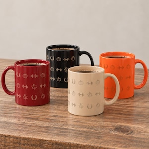 May include: Four ceramic mugs in red, black, cream, and orange, each featuring a repeating pattern of etched icons including pumpkins, maple leaves, and horseshoes. Each mug has a side handle and contains a dark liquid, arranged on a wooden surface.