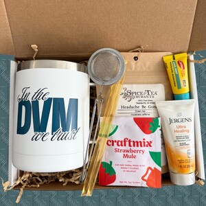 May include: A gift box containing a white tumbler with "In the DVM we trust" text, a tea infuser, tea, lip balm, a packet of strawberry mule mix, and a tube of Jergens Ultra Healing lotion. The box is filled with crinkle paper.