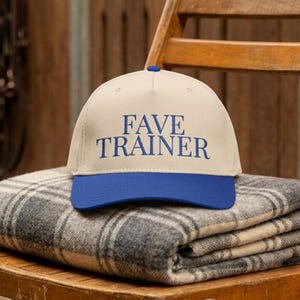 May include: A beige baseball cap with a blue brim and the text "FAVE TRAINER" in blue. The hat sits on a folded gray and white plaid blanket, placed on a wooden chair.