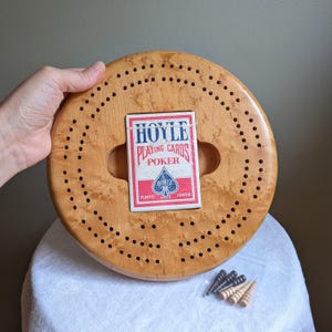 May include: A wooden cribbage board with a circular design and numerous small holes. A deck of Hoyle Playing Cards is placed in the center. Includes a set of cribbage pegs. The board is made of light-colored wood.