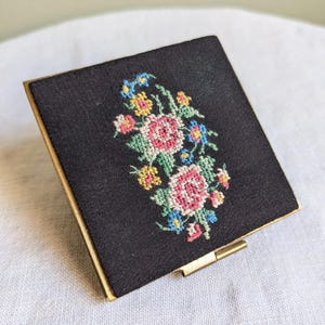 May include: A vintage black compact mirror with a floral design embroidered on the front. The flowers are pink, yellow, blue, and green.