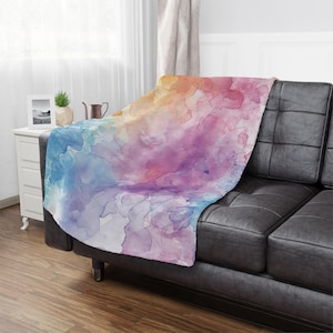 May include: A soft, plush throw blanket with a watercolor design featuring shades of blue, purple, pink, and orange. The blanket is draped over a black leather couch.